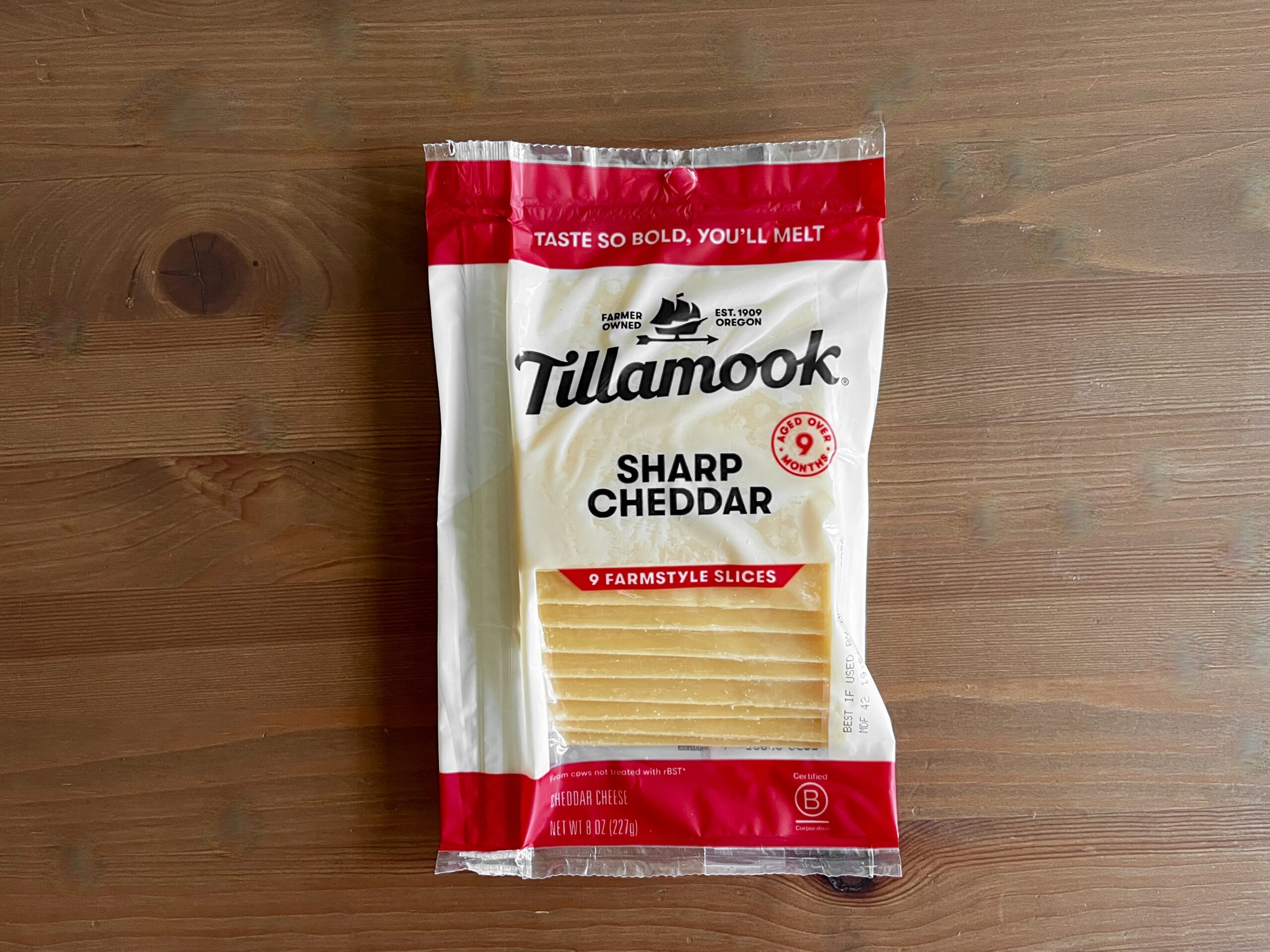 A package of Tillamook Sharp Cheddar Farmstyle Slices sits on a wooden surface. The packaging is white and red, featuring branding and a clear window showing the cheese slices inside.