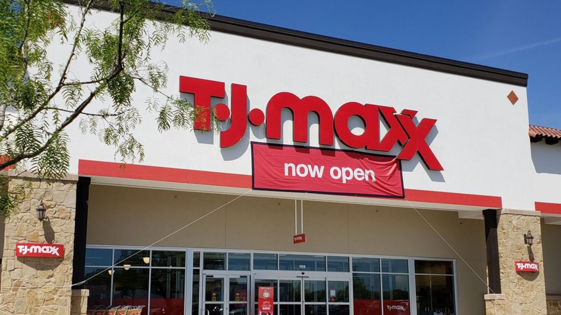 The front entrance of a TJ Maxx store with a large red sign and a banner underneath that reads "now open" on the white building facade.