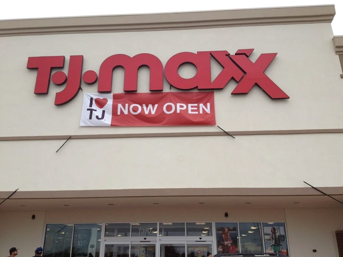 The exterior of a TJ Maxx store with a large red logo on the building and a banner that says "I ♥ TJ NOW OPEN" hanging below the sign above the entrance.
