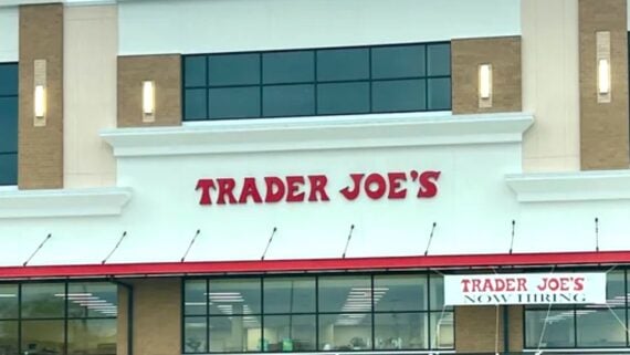 The front of a Trader Joe’s store with large red letters spelling "TRADER JOE'S" and a banner below that reads "NOW HIRING." The building has large windows and beige accents.