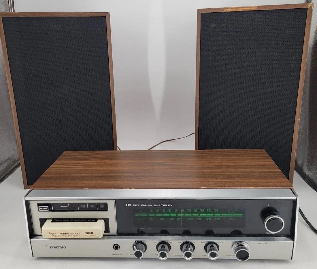 A vintage Bradford stereo receiver with a faux wood top, silver front panel, multiple knobs, and buttons, sits in front of two black rectangular speakers. An 8-track tape is inserted into the player.