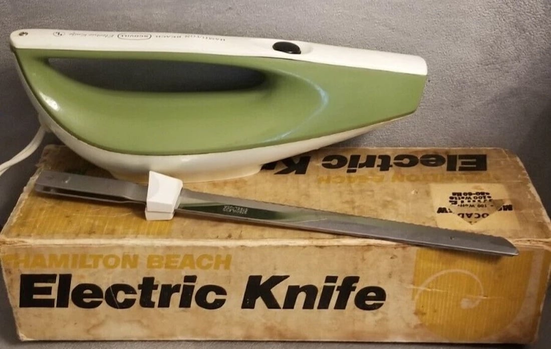A vintage green and white Hamilton Beach electric knife rests on its original box, with a detachable serrated blade placed in front of the box. The box features bold black and yellow text.