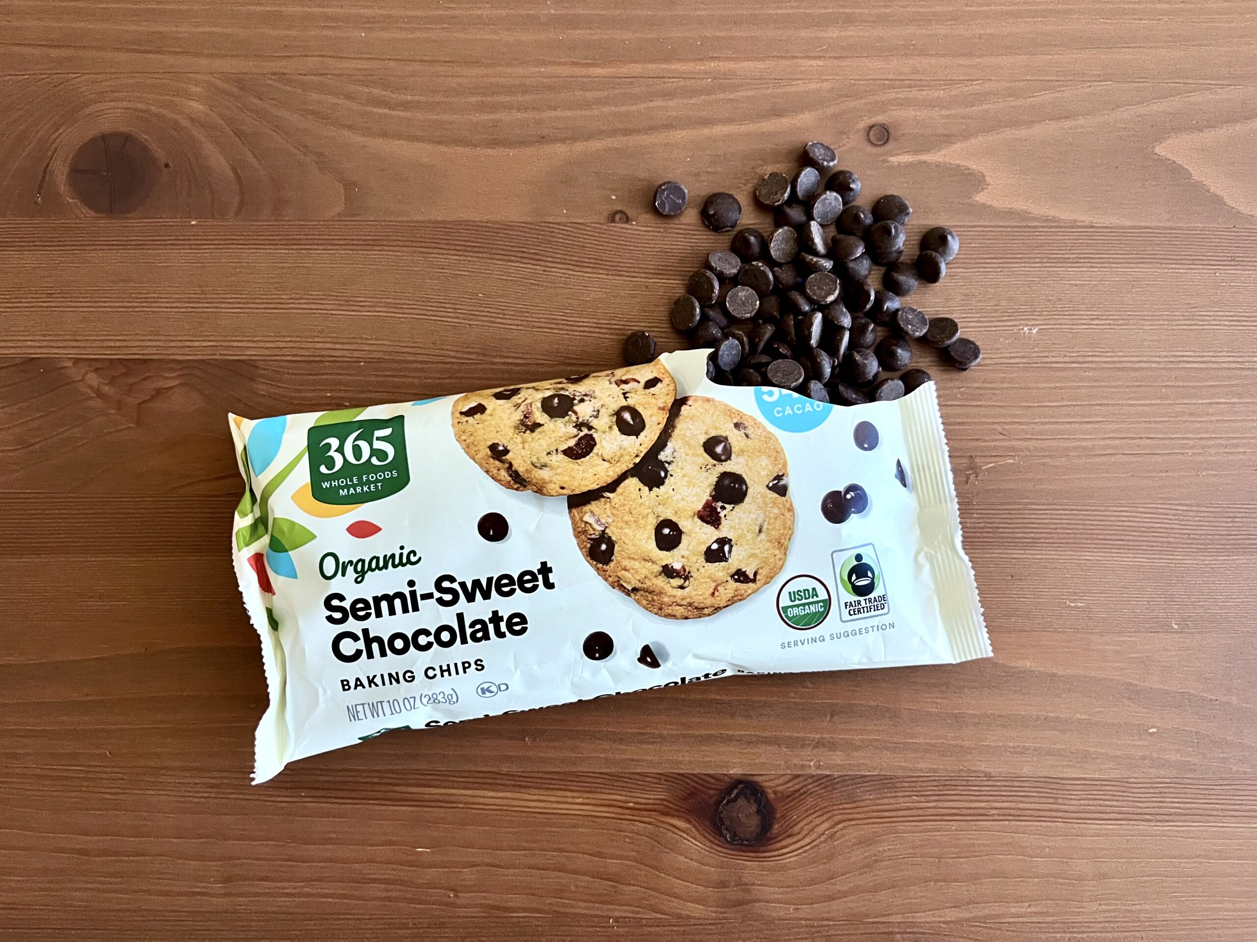 A package of 365 Organic Semi-Sweet Chocolate Baking Chips sits on a wooden surface, with some chocolate chips scattered next to it.