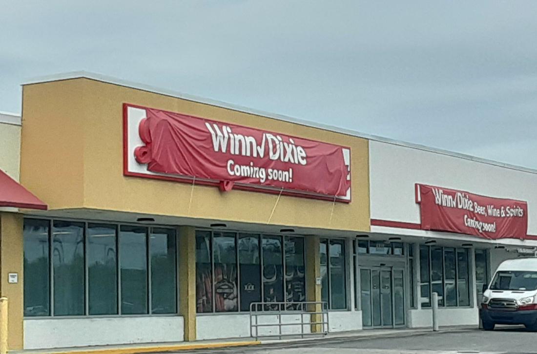 A storefront with a large red banner that reads "Winn/Dixie Coming soon!" is displayed above the entrance. The building appears unoccupied, and the windows are empty.