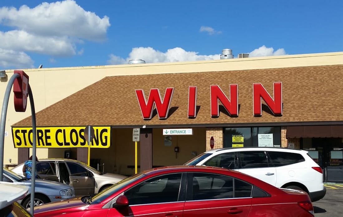 A store with a large red "WINN" sign has a yellow "Store Closing" banner across the entrance. Several cars are parked in front under a sunny blue sky with scattered clouds.