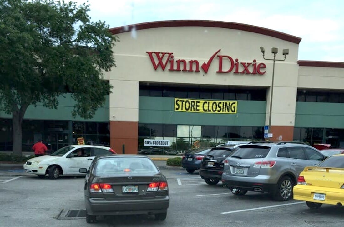 A Winn-Dixie grocery store with a large yellow "STORE CLOSING" sign above the entrance. Several cars are parked in front of the store on a cloudy day.