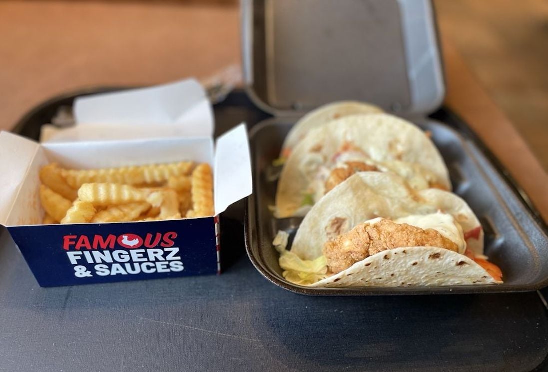 A tray with a box of crinkle-cut French fries labeled “Famous Fingerz & Sauces” next to an open container holding three soft tacos filled with fried chicken, lettuce, and tomato.