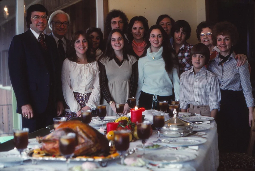 A group of thirteen people, spanning multiple generations, stand and smile behind a dining table set for a meal, featuring a roasted turkey, candles, and drinks. The setting appears festive and familial.