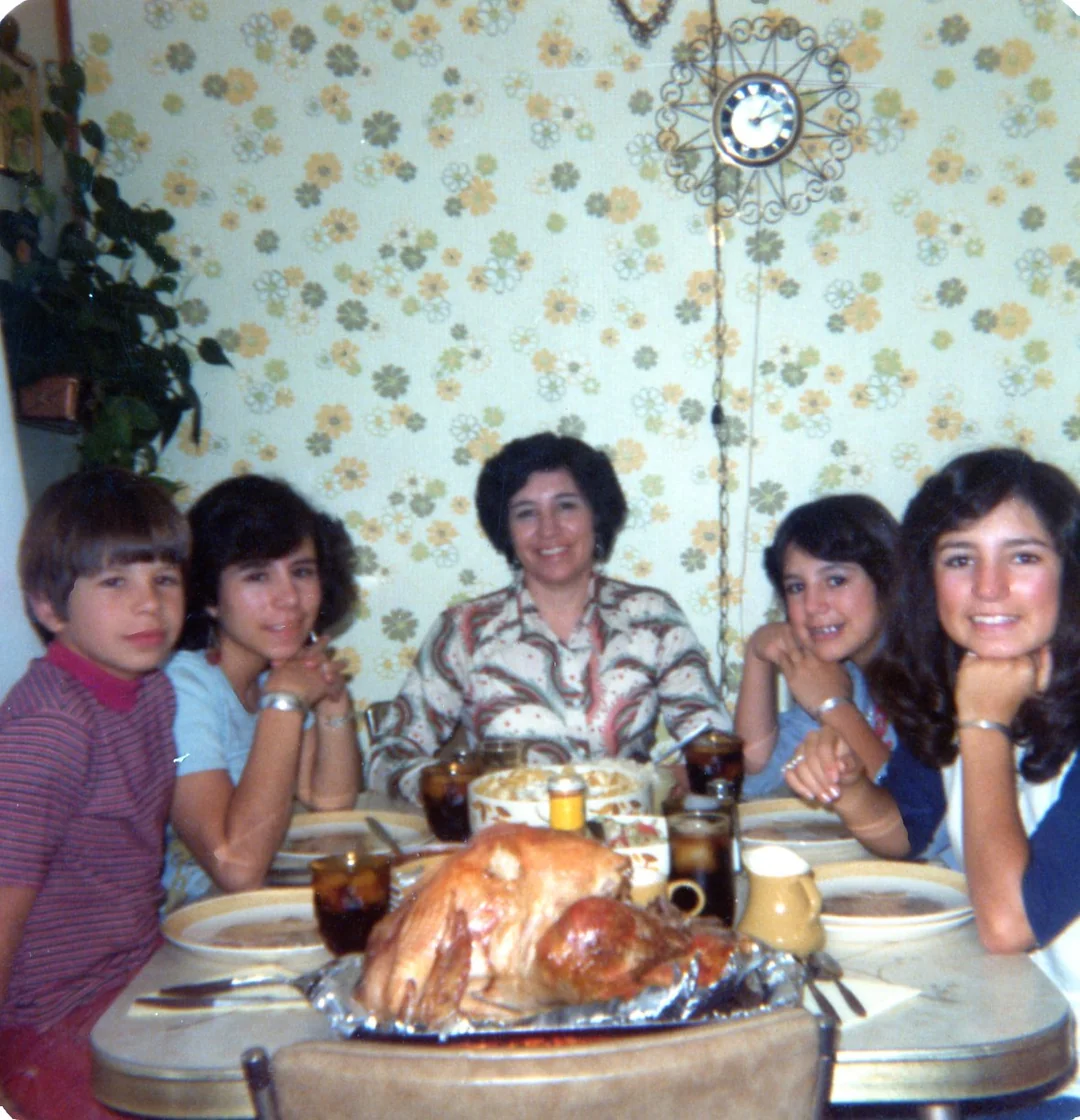 Five people sit smiling at a dining table set for a meal, with a roast turkey in the center. The background shows floral wallpaper, a hanging plant, and a clock on the wall.