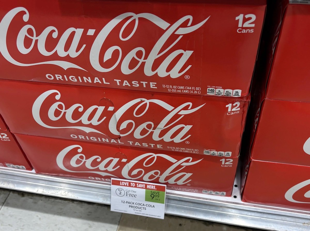 Red cases of Coca-Cola Original Taste, each containing 12 cans, are stacked on a store shelf. A sale sign in front advertises a "Buy 2, Get 1 Free" offer on 12-pack Coca-Cola products.