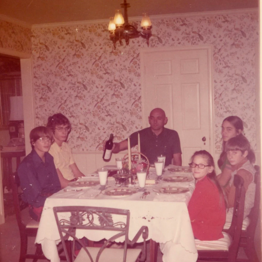 A family of six sits around a dining table set for a meal. The man at the head of the table holds a bottle. The room has floral wallpaper and a chandelier overhead. Everyone looks toward the camera.