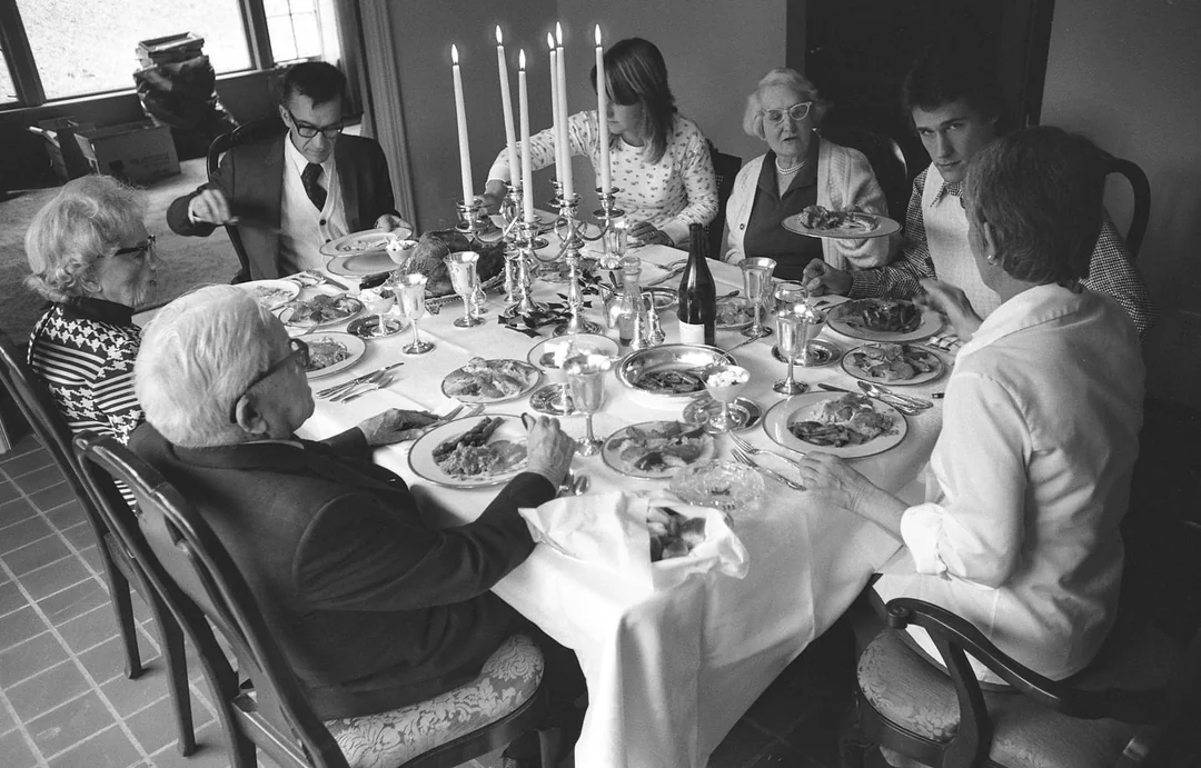 A black and white photo of seven people sitting around a dining table set with candles, plates of food, and silverware, sharing a meal together in a warmly-lit room captures the charm of vintage Thanksgiving photos.