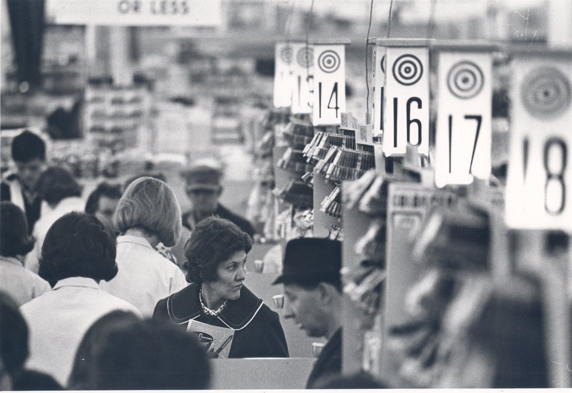 Black-and-white photo of shoppers standing in line at supermarket checkout lanes, each marked with numbered signs, including 14, 16, 17, and 18. People appear to be waiting to pay for their groceries.