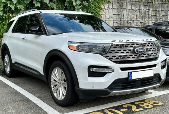 A white Ford Explorer SUV is parked in a parking space beside a stone wall with greenery above. The vehicle has tinted windows and chrome details.