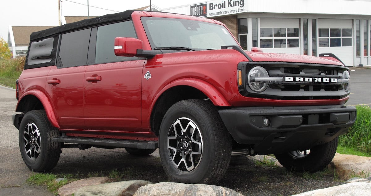A red Ford Bronco SUV is parked outside, with raindrops visible on its surface. It has a black roof, rugged tires, and is positioned on a slight incline in front of a building.
