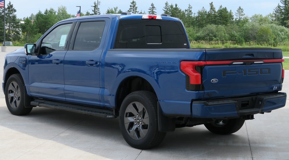 A blue Ford F-150 pickup truck is parked on a concrete surface, viewed from the rear side. Trees and greenery are visible in the background under a partly cloudy sky.