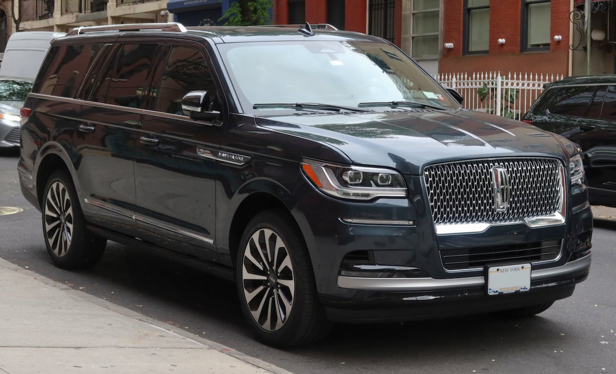 A dark-colored Lincoln Navigator SUV is parked on a city street near a sidewalk, with buildings and trees visible in the background.