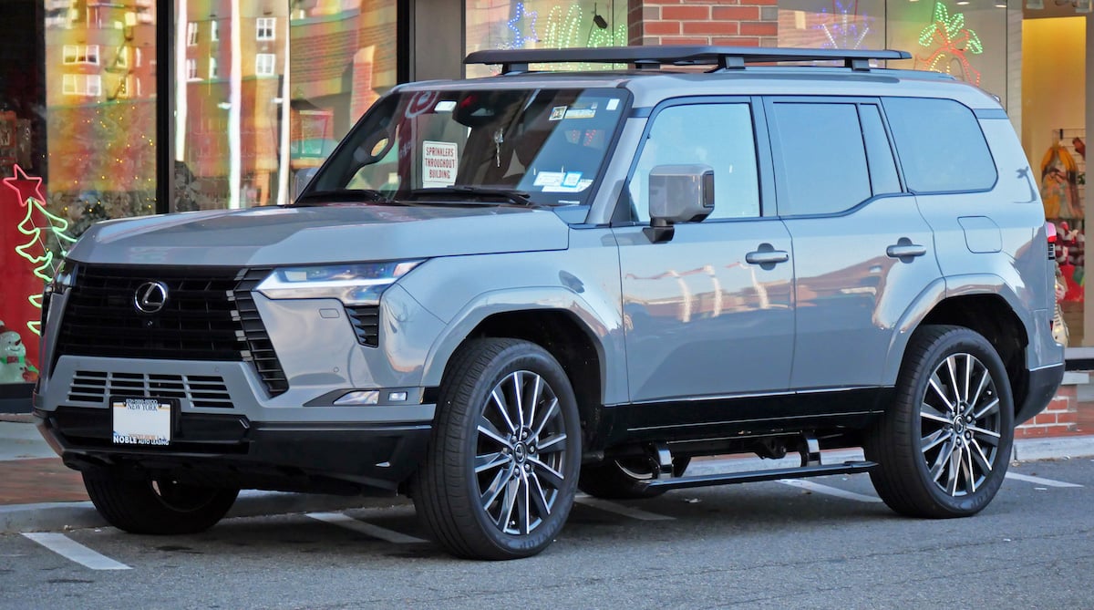 A gray Lexus SUV is parked on the street in front of a store with holiday window decorations. The vehicle has large black alloy wheels and black roof rails.