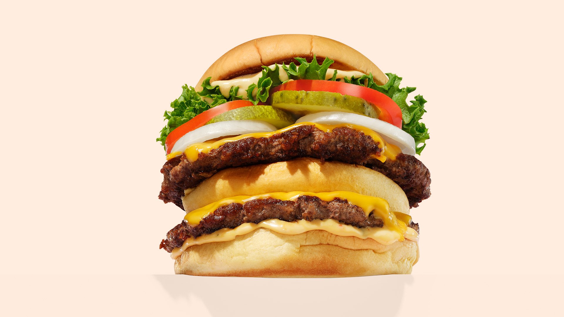 A double cheeseburger with two beef patties, melted cheese, lettuce, tomato, pickles, onion, and sauce in a soft bun, shown against a plain light background.