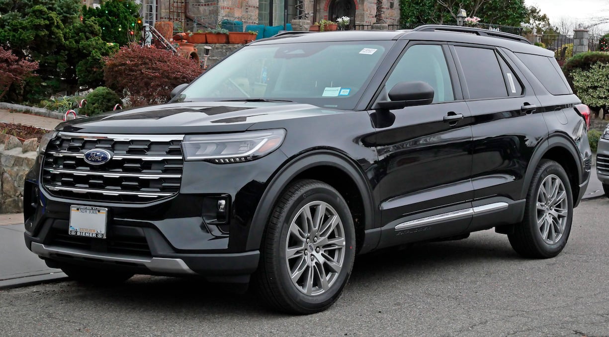 A black Ford Explorer SUV is parked on a residential street near a stone wall and greenery, with houses visible in the background.
