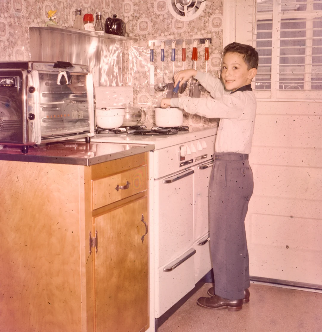 A young boy smiles while standing at a vintage stove, stirring a pot in a retro kitchen with patterned wallpaper, a toaster oven, and utensils hanging on the wall.