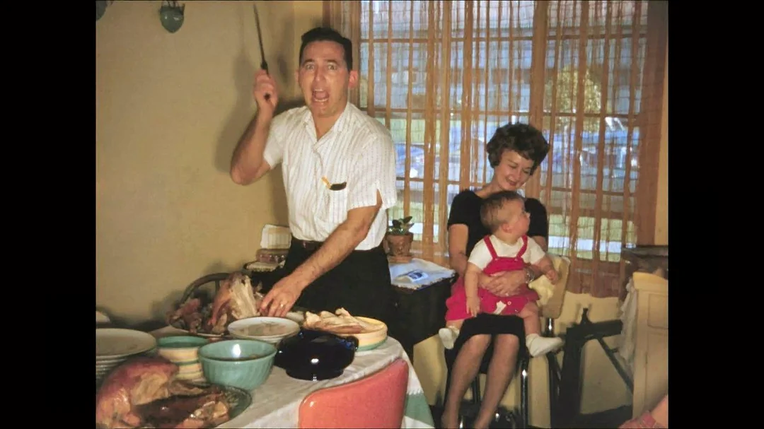 A man enthusiastically carves a turkey at a dining table filled with dishes, while a woman sits nearby holding a baby on her lap. Sunlight streams through the window behind them.