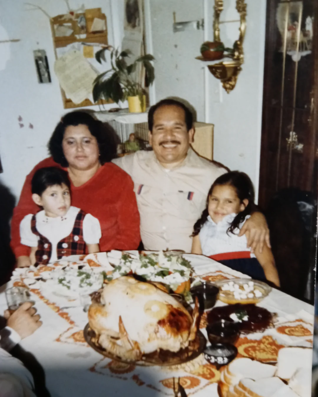 A family of four sits at a dining table with a large roasted turkey and other dishes. The parents are smiling, their two daughters beside them. The room has warm decor with plants, photos, and wall hangings in the background.
