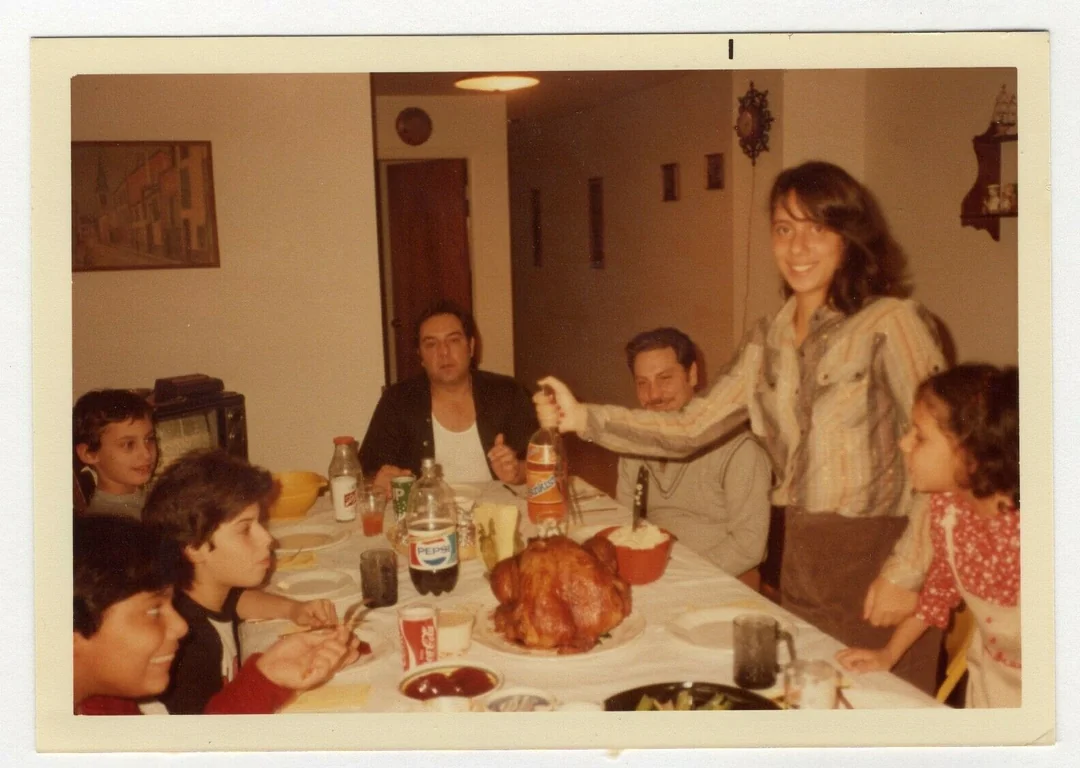 A group of people sit around a dining table set with food, including a roast turkey. A smiling woman stands by the table holding a condiment, while a young girl stands next to her. The scene appears to be a family meal.