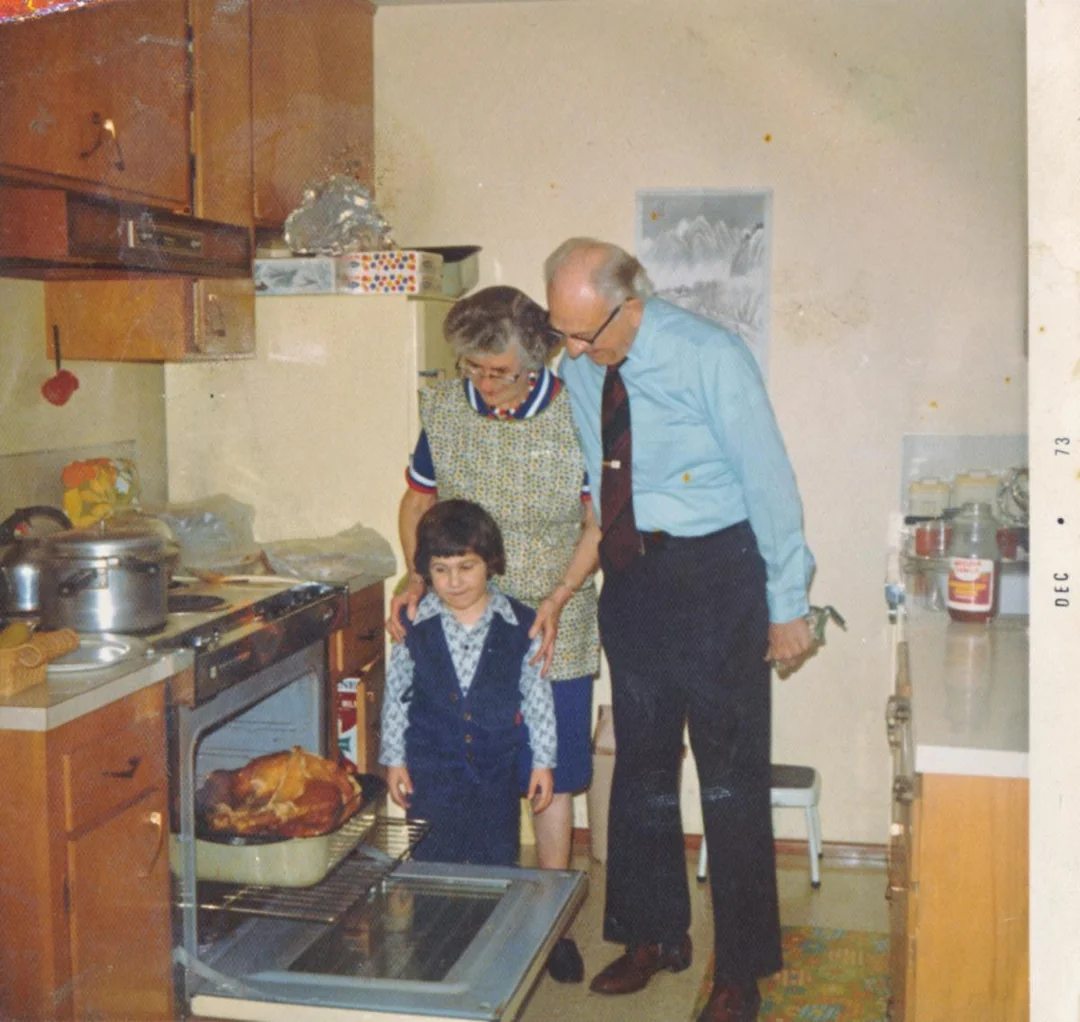 An elderly couple and a young child stand in a kitchen, looking at a roasted turkey in the oven. The woman wears an apron, the man wears a tie, and the child is in a blue vest. Pots and kitchen items are visible around them.