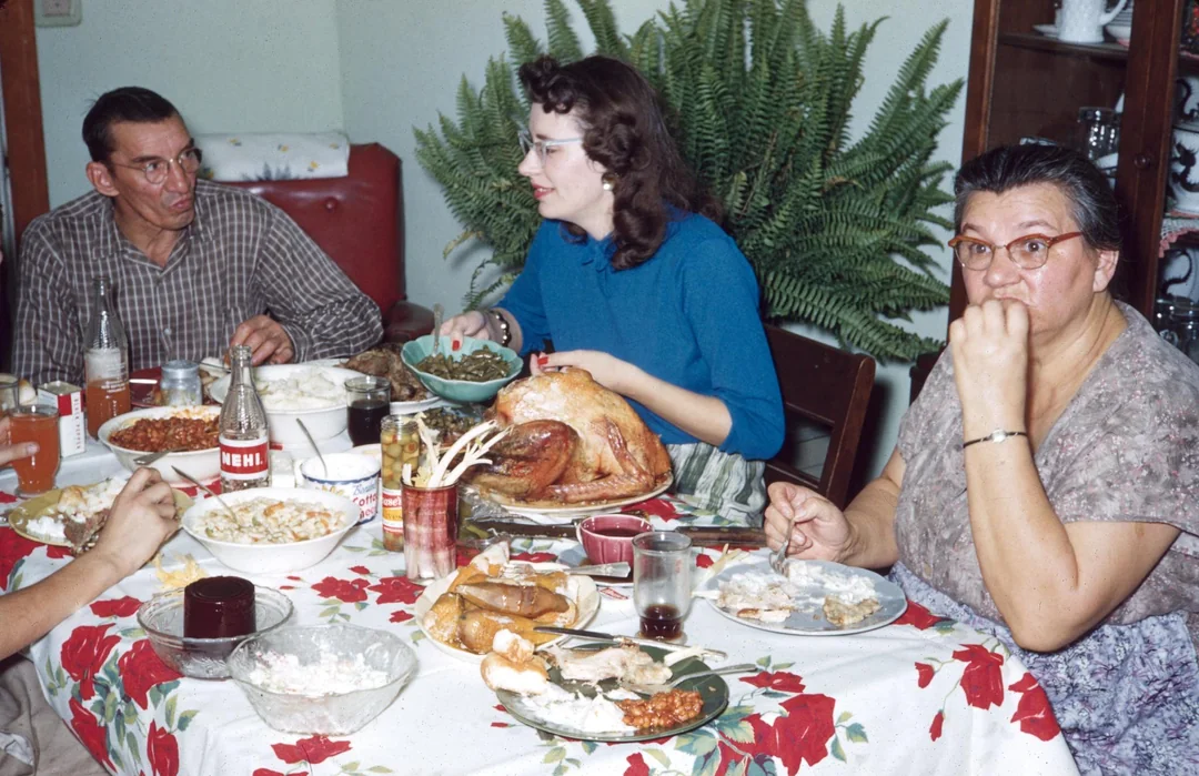 Three adults sit around a dinner table with a floral tablecloth, sharing a meal of turkey, green beans, and sides. Capturing the warmth of vintage Thanksgiving photos, one woman looks at the camera as others converse; a fern plant is in the background.