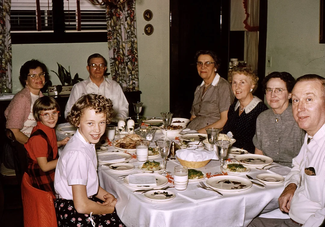 Seven people, including children and adults, sit around a dining table set with plates, glasses, and food. Smiling and enjoying a meal together in a warmly lit, homey dining room, the scene recalls vintage Thanksgiving photos.