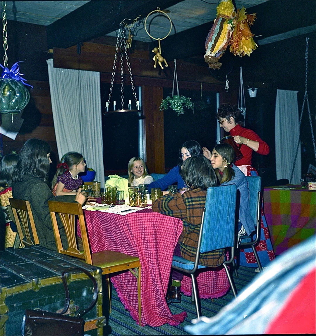 A group of children sit around a table covered with a red checkered cloth at a cozy indoor party. An adult stands nearby. Decorations, including a piñata, hang from the ceiling. The room is warmly lit and festive.