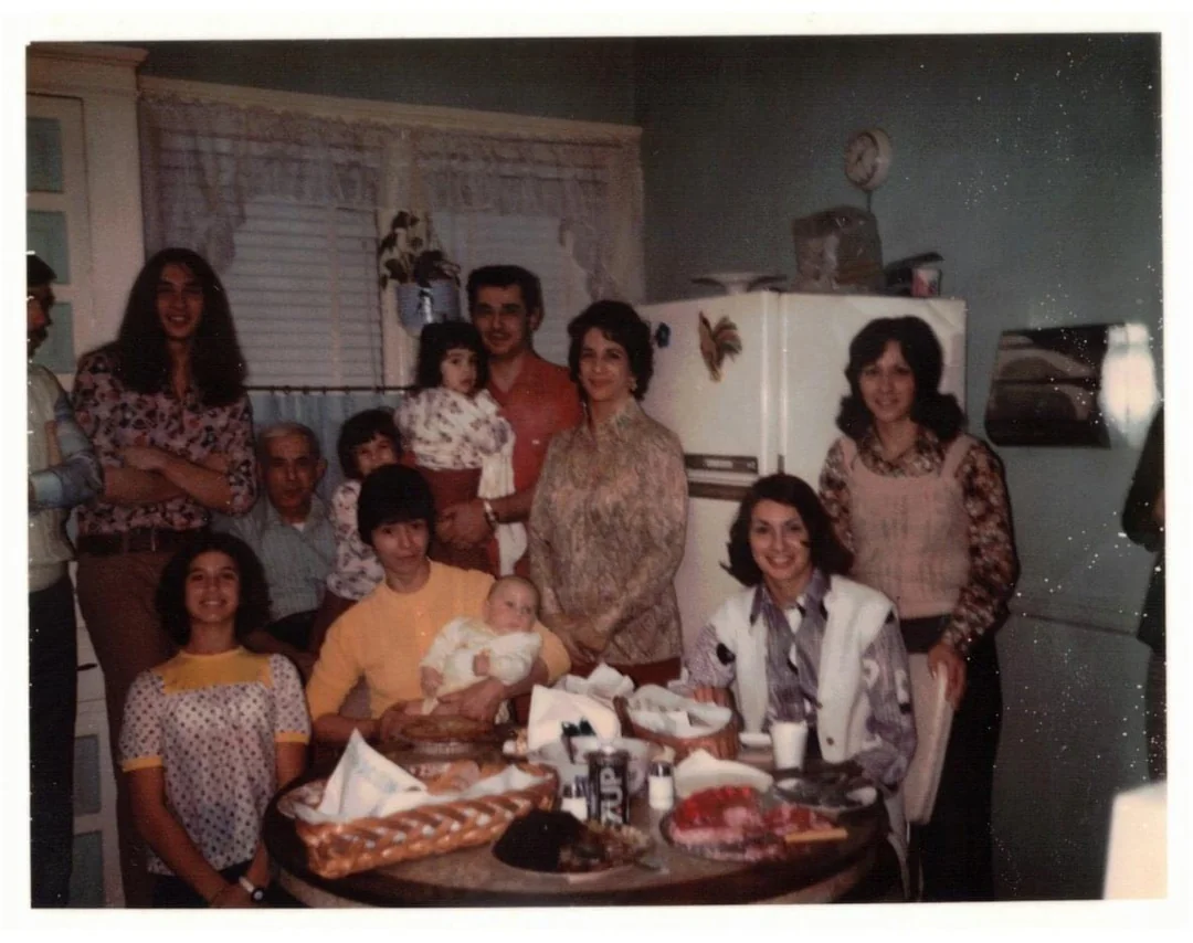 A vintage photo of a smiling family gathered in a kitchen around a table with food. Three children and several adults pose together, with a refrigerator and window in the background. The scene feels warm and nostalgic.