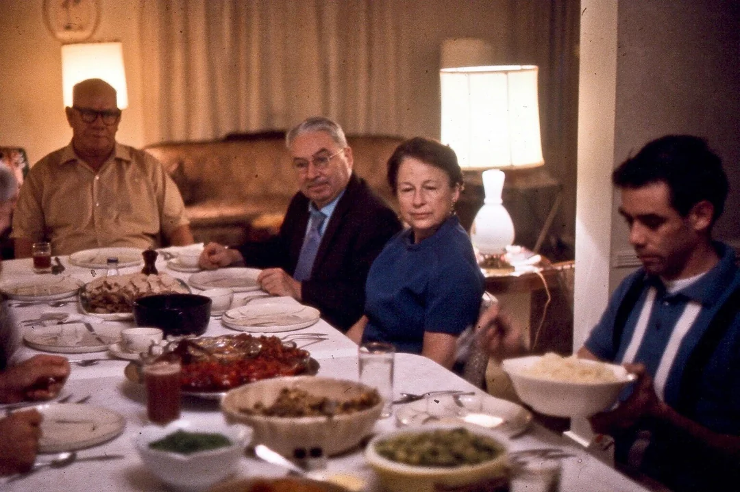 Four people sit around a dining table set with various dishes, including salads and meats. The setting appears to be a cozy living room with warm lighting from a lamp in the background.