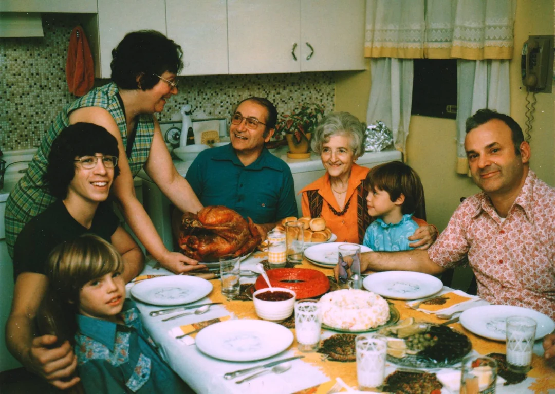 A family of seven, including children and seniors, sits around a dining table in a kitchen. A woman serves a roast turkey, while everyone smiles, creating a warm, festive atmosphere. Food and drinks fill the table.