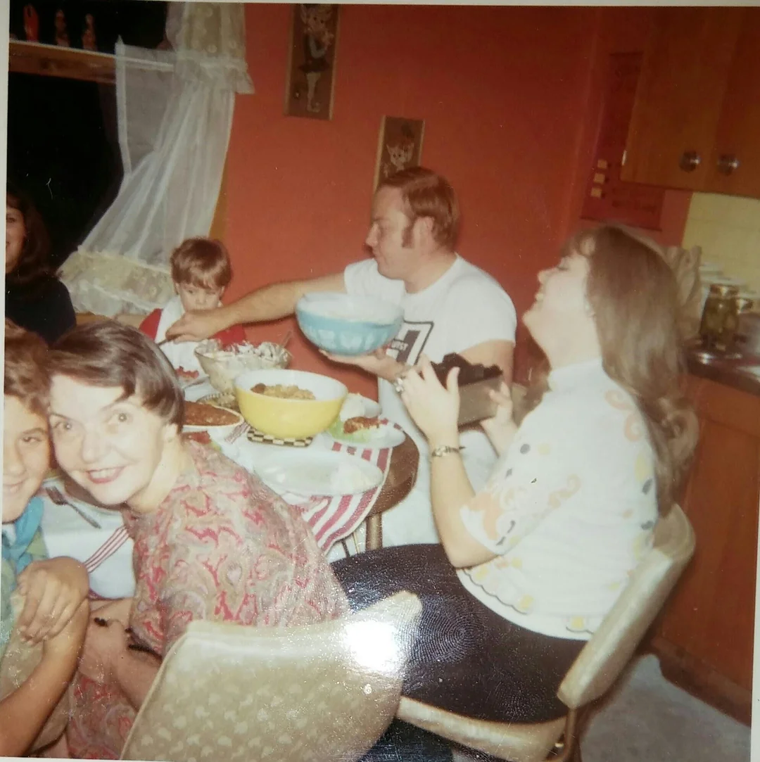 A group of people sit around a dining table in a cozy kitchen, sharing a meal and laughing. Bowls of food are on the table. A woman in a patterned shirt smiles at the camera, while others are engaged in conversation.