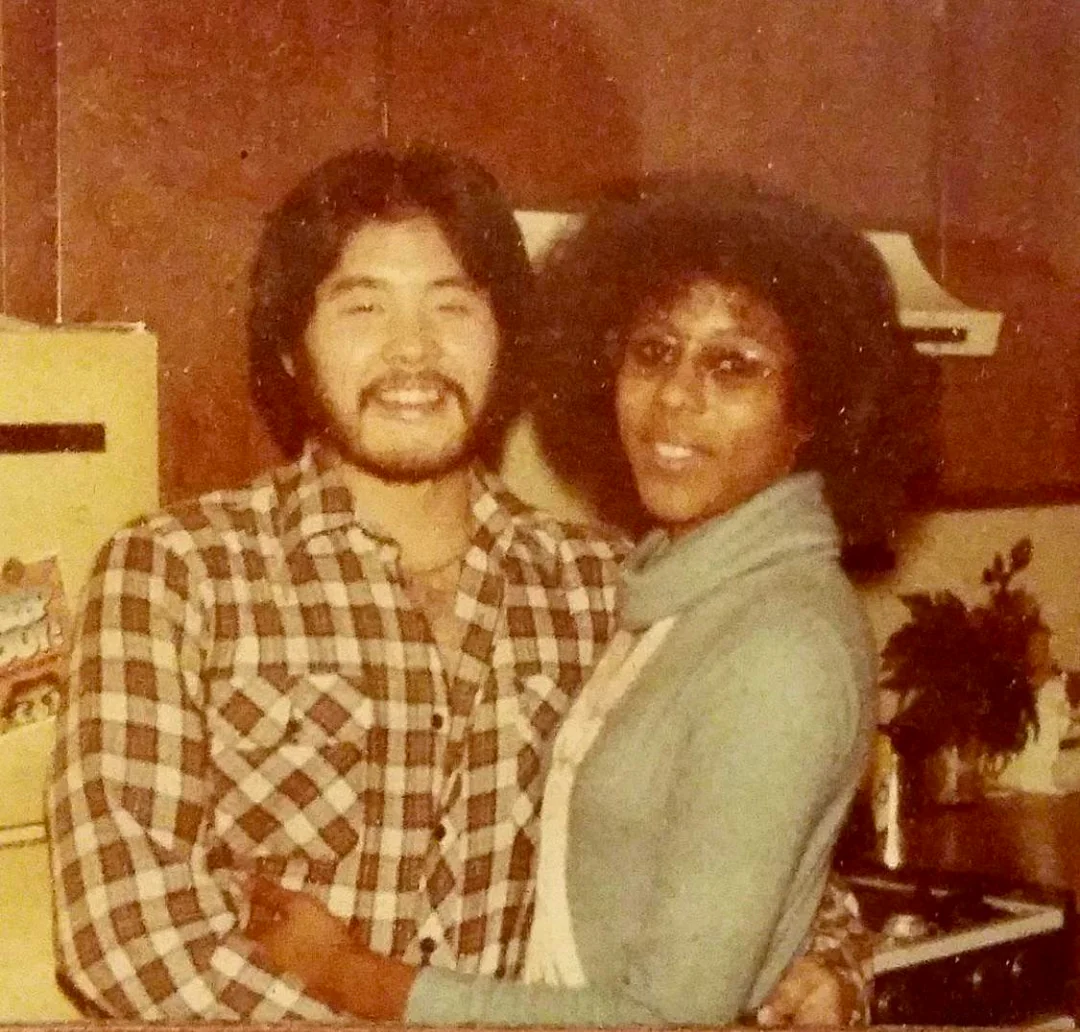 A man in a plaid shirt and a woman in glasses and a light blue sweater smile and pose together in a kitchen with wooden cabinets and a yellow refrigerator in the background.