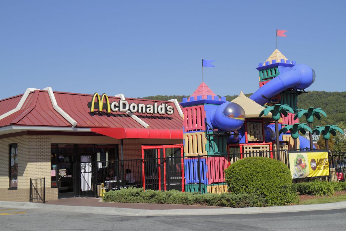 A McDonald's restaurant with a colorful outdoor playground featuring slides and climbing structures, set against a clear blue sky and green, wooded hills in the background.