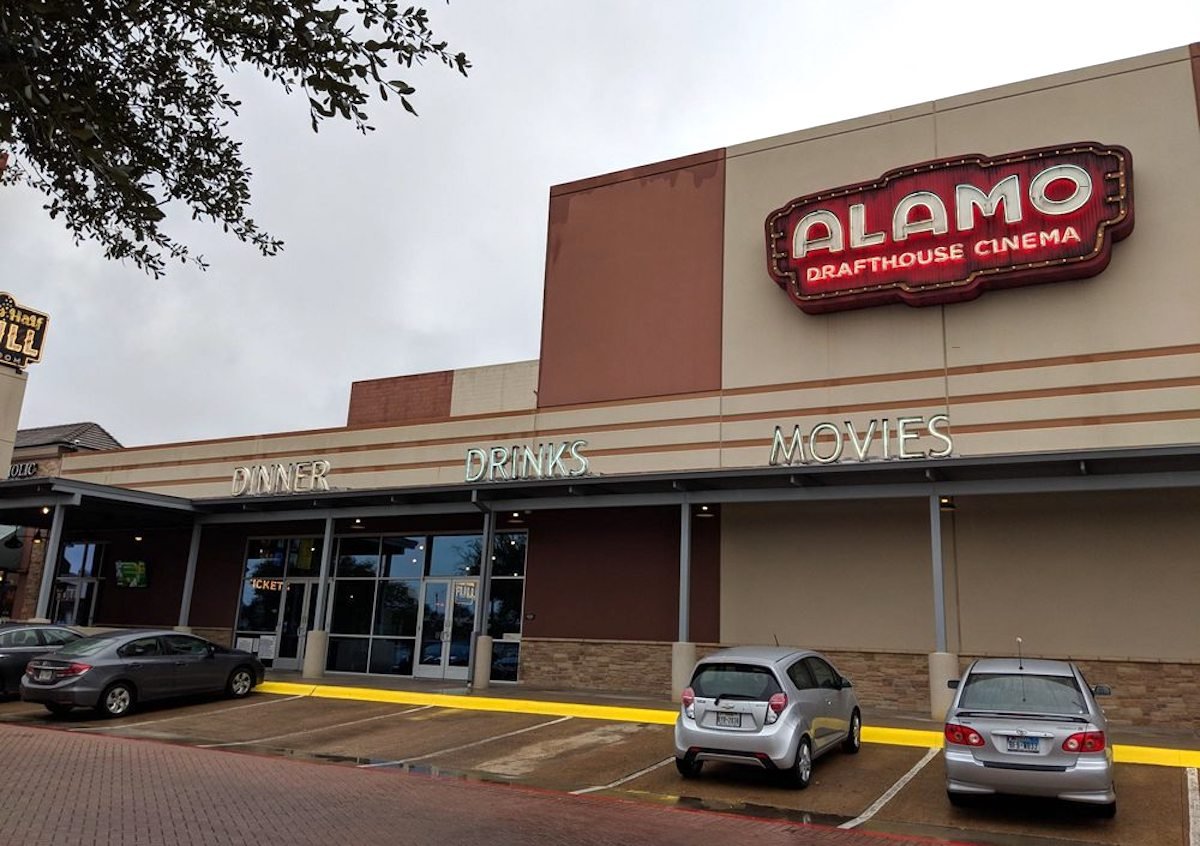 Alamo Drafthouse Cinema building with a large sign above the entrance. Neon words "Dinner," "Drinks," and "Movies" are displayed above the windows. Three cars are parked in front of the cinema on a cloudy day.