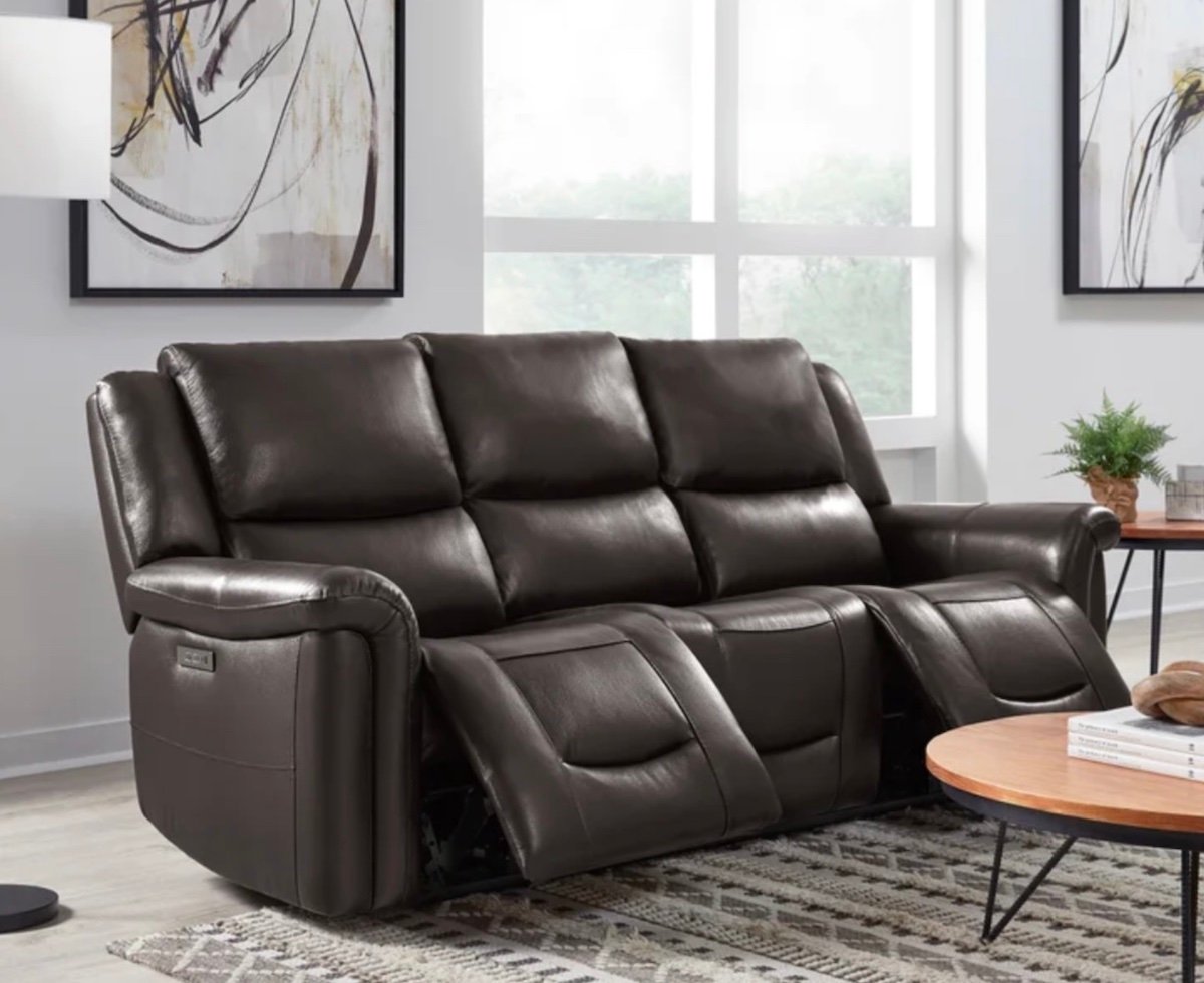 A modern living room features a dark brown leather reclining sofa with two extended footrests, a round wooden coffee table, a potted plant, abstract wall art, and large windows letting in natural light.