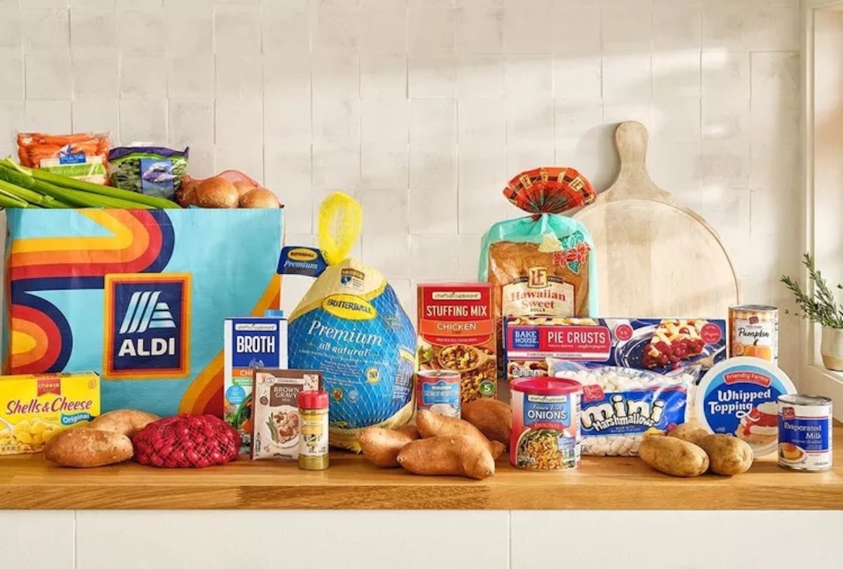 A variety of groceries, including fresh vegetables, a turkey, canned goods, spices, and baking ingredients, are arranged on a wooden counter next to an ALDI shopping bag in a bright kitchen.