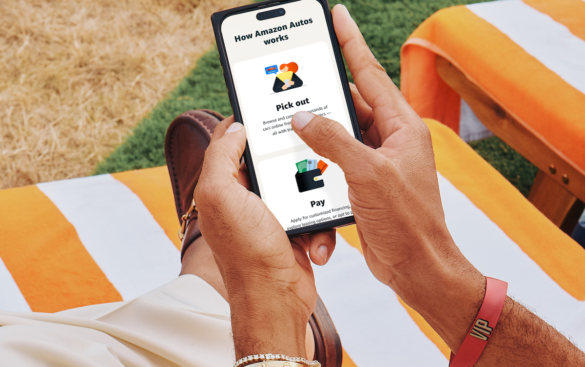 A person holding a smartphone reads "How Amazon Autos works" on the screen while sitting on an orange-and-white striped lounge chair outdoors.