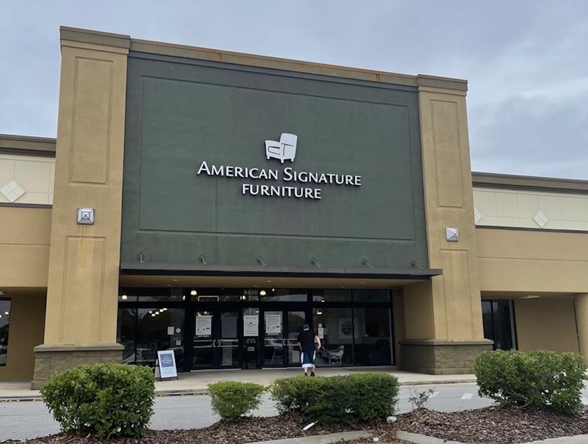 The front of an American Signature Furniture store with large signage, glass entrance doors, and a person standing outside. Bushes and a parking lot are visible in the foreground under a cloudy sky.