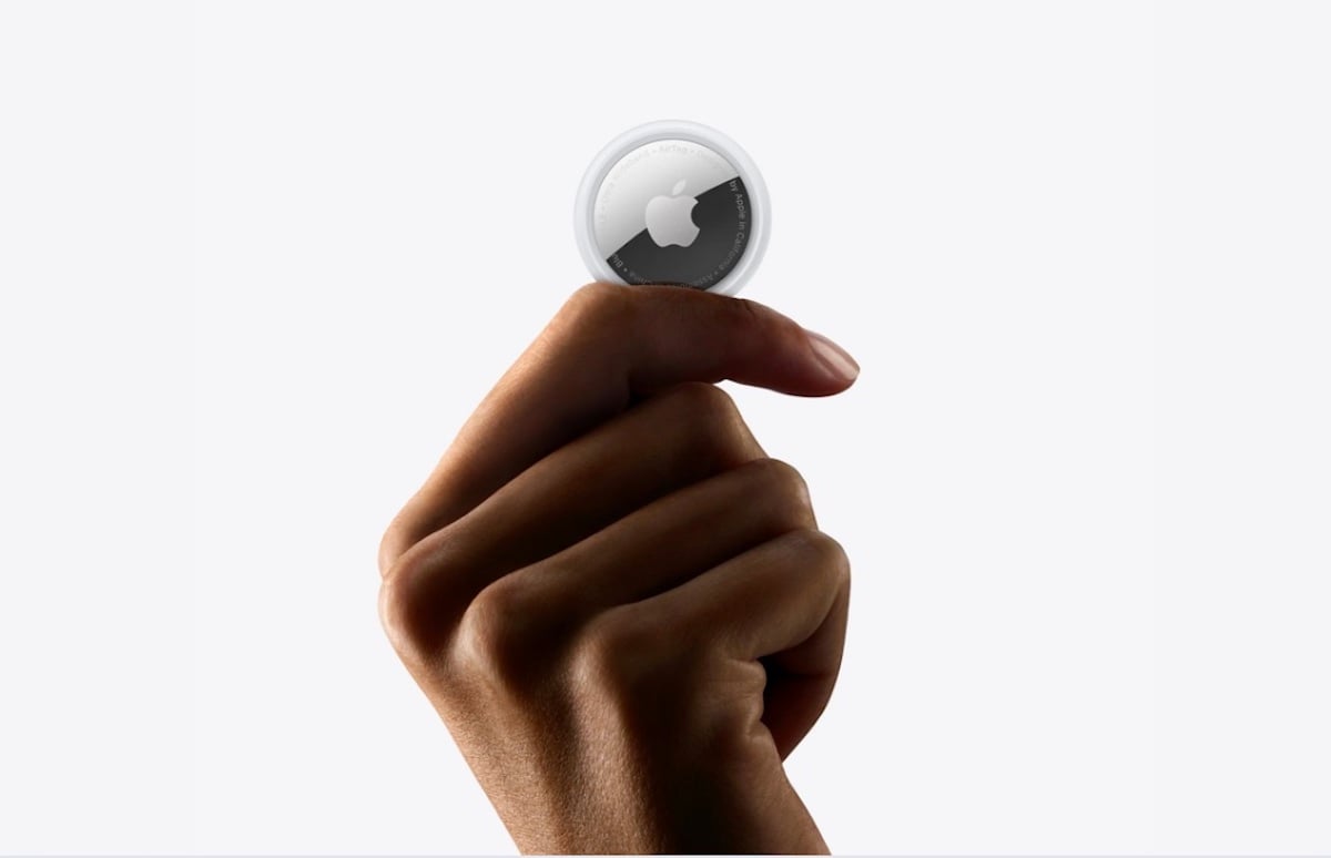 A hand holding an Apple AirTag between the thumb and index finger against a plain white background.