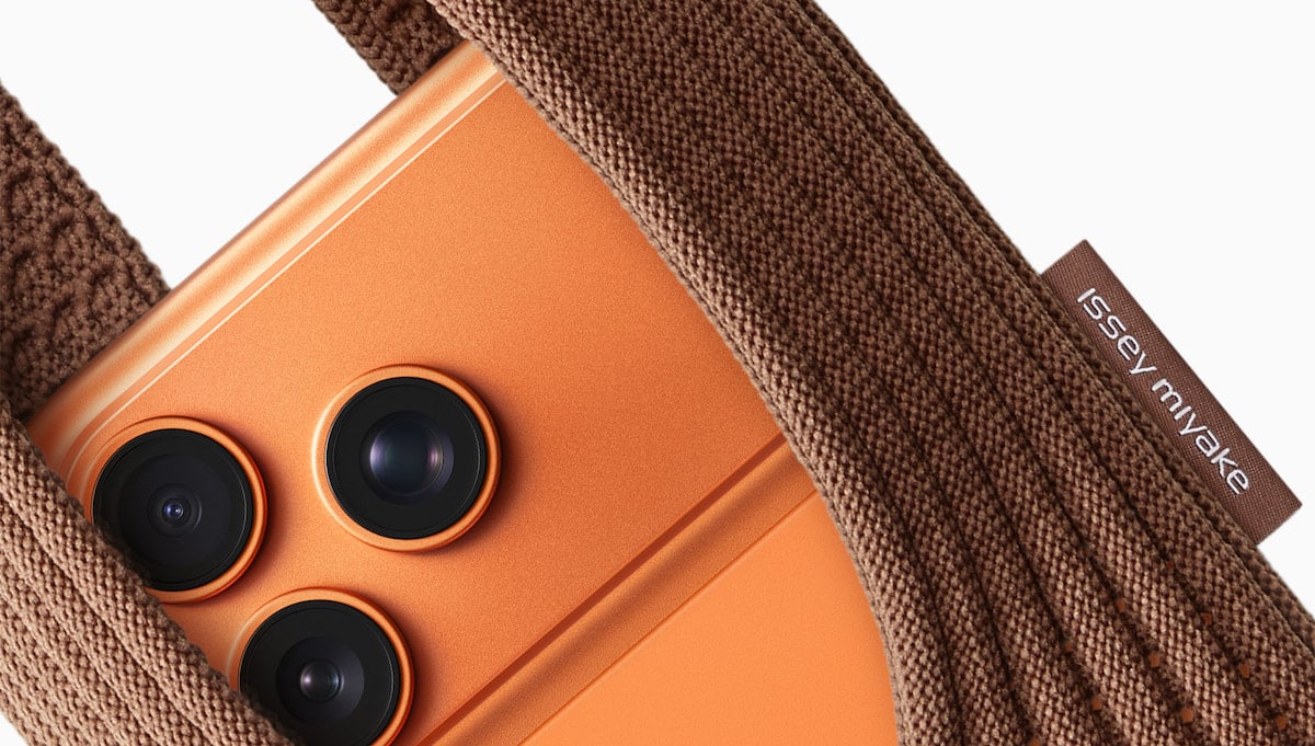 A close-up of an orange smartphone with three camera lenses, partially tucked into a textured brown fabric pouch with a tag that reads "JESSE MIN XE" on the side.