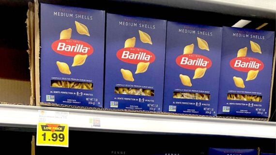 Four boxes of Barilla Medium Shells pasta are displayed on a grocery store shelf with a yellow price tag below showing "Low Price $1.99". The boxes are blue with yellow pasta shell images.