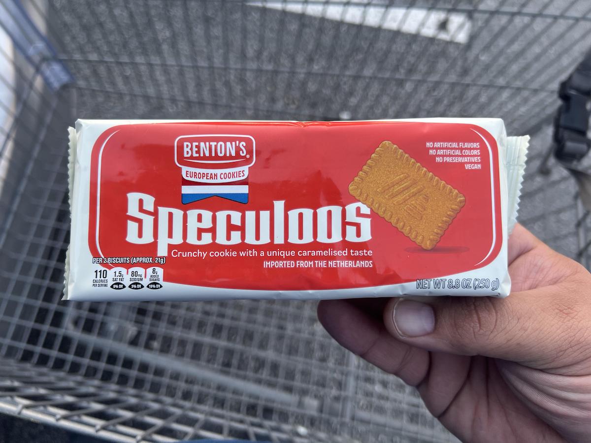 A hand holds a package of Benton's Speculoos European Cookies in front of a shopping cart. The package is red and white, showing an image of the cookie and product details.