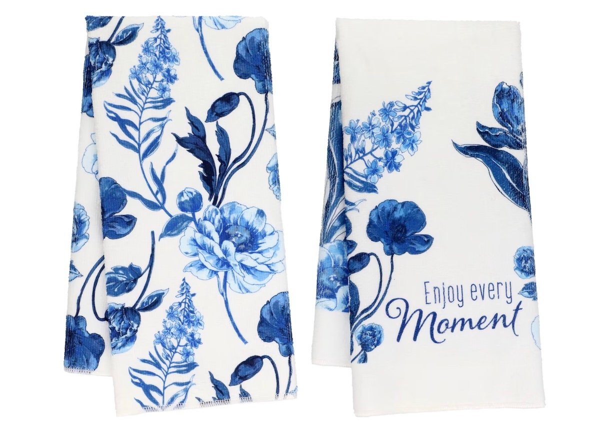 Two white kitchen towels with blue floral designs; one has only flowers, the other features flowers and the phrase “Enjoy every Moment” written in blue script.