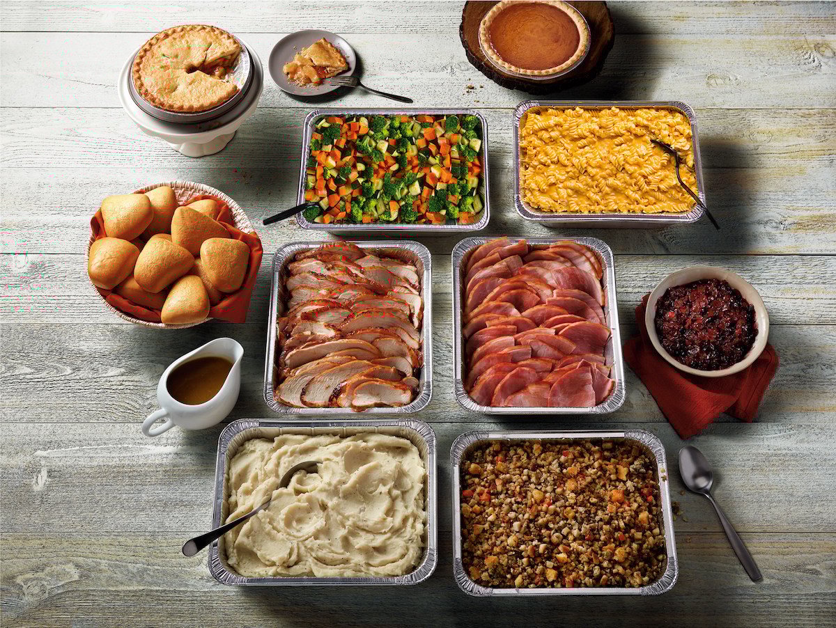 A variety of Thanksgiving foods on a table, including sliced turkey, ham, stuffing, mashed potatoes, gravy, dinner rolls, mixed vegetables, cranberry sauce, sweet potato casserole, and two pies.