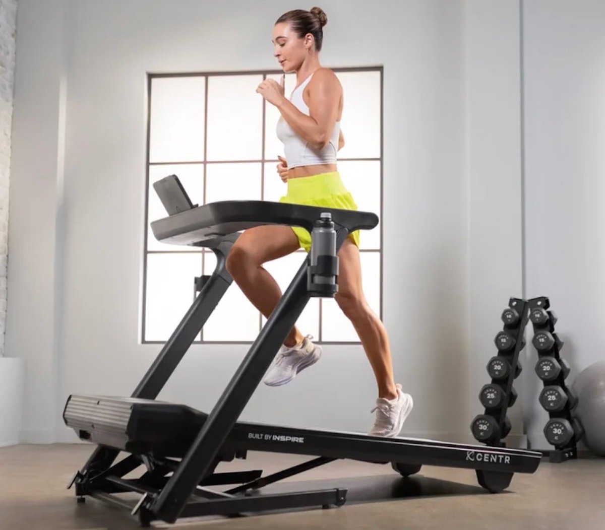 A woman in a white top and yellow shorts runs on a treadmill in a bright gym room. There is a rack of dumbbells and a water bottle on the treadmill. Sunlight streams through a large window in the background.